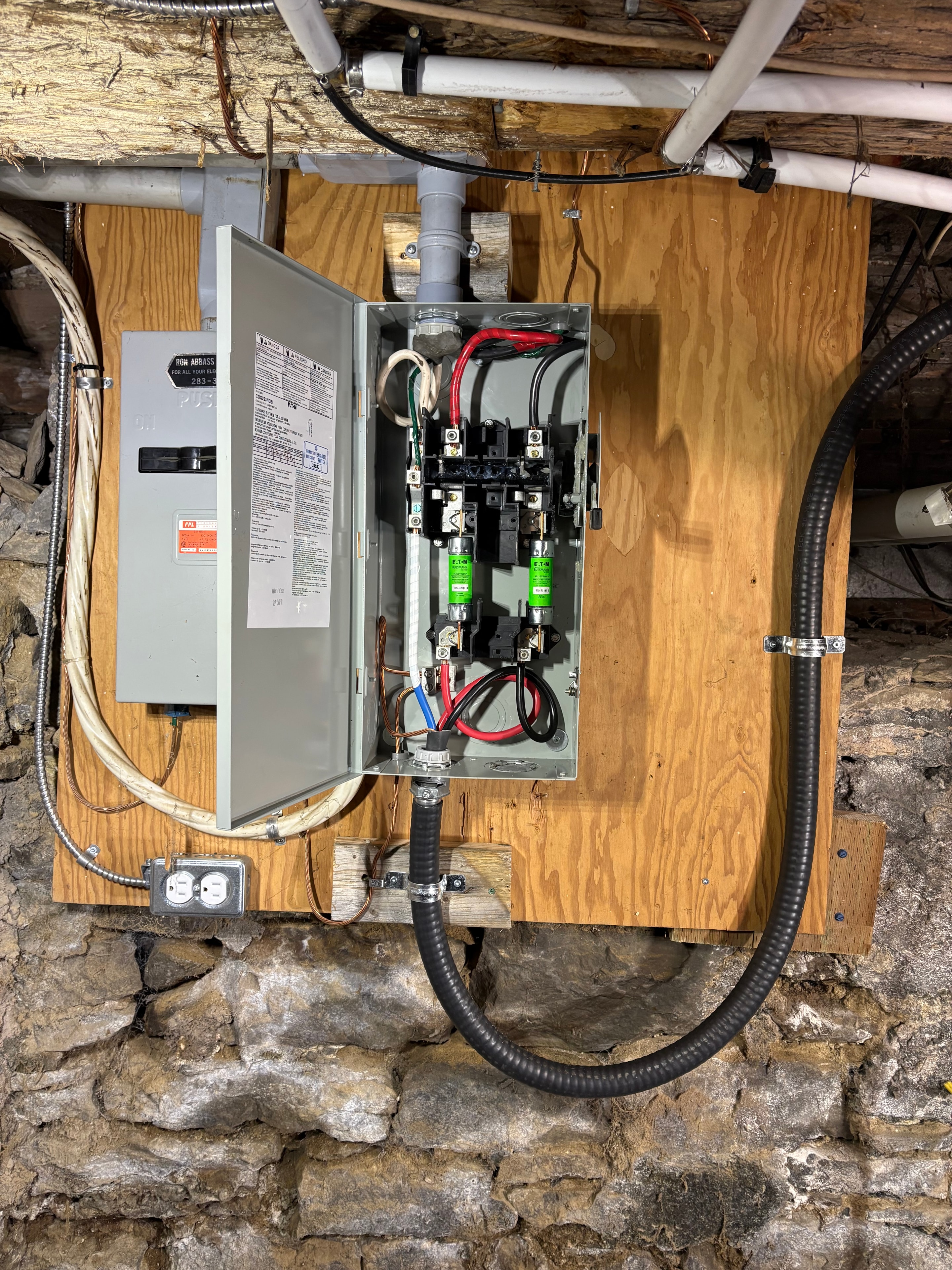 New electrical service panel open showing main breaker, branch-circuit connections and flexible conduit runs, mounted on plywood against a stone foundation wall